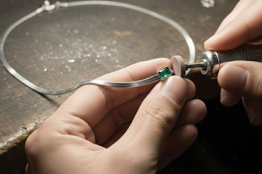 A close-up of an artisan hand-polishing a bezel-set emerald on a silver herringbone chain, illustrating the detailed hand-finishing process of Montkai jewelry.